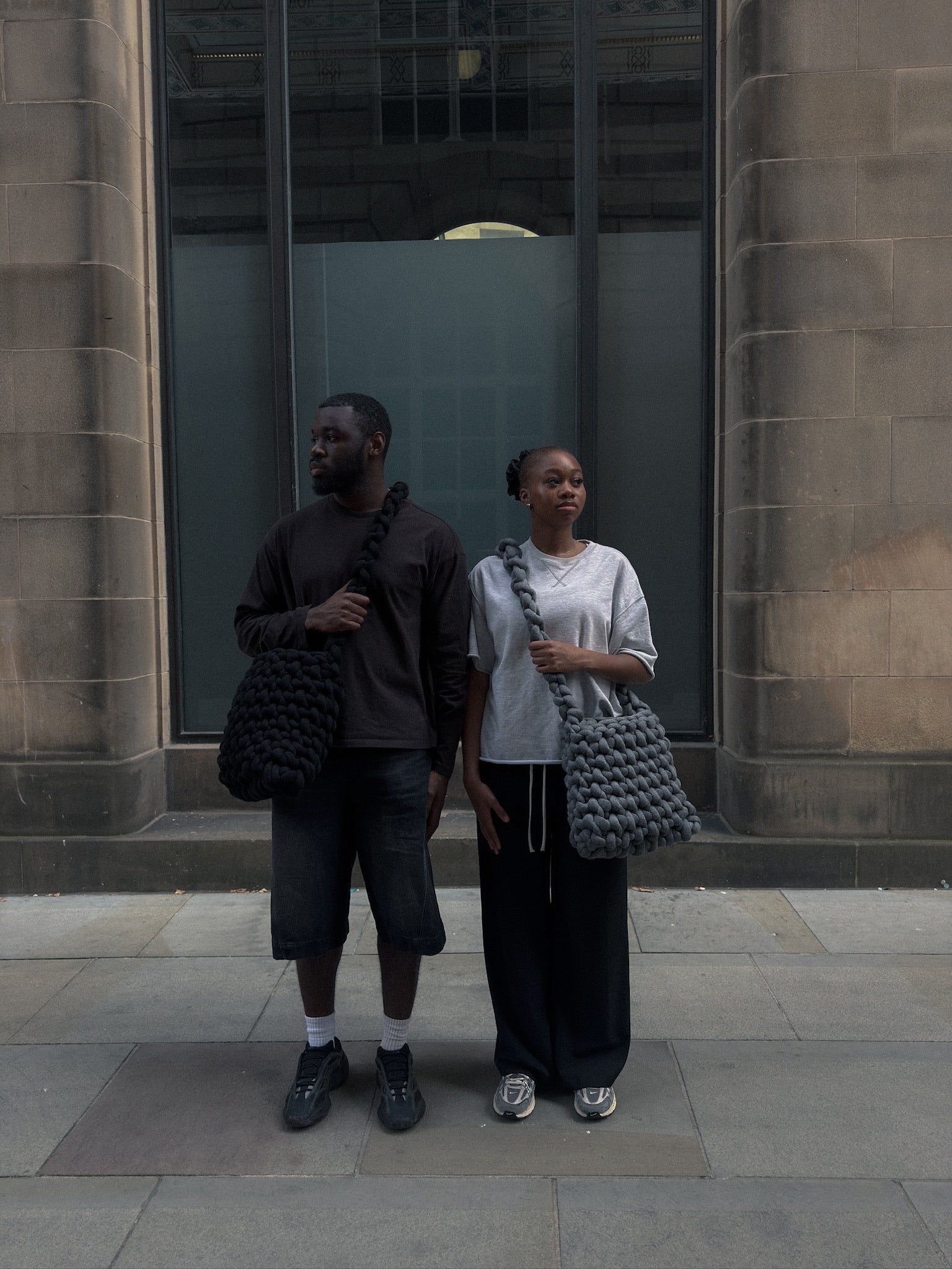 Two people standing in front of a building, each holding a textured bag.