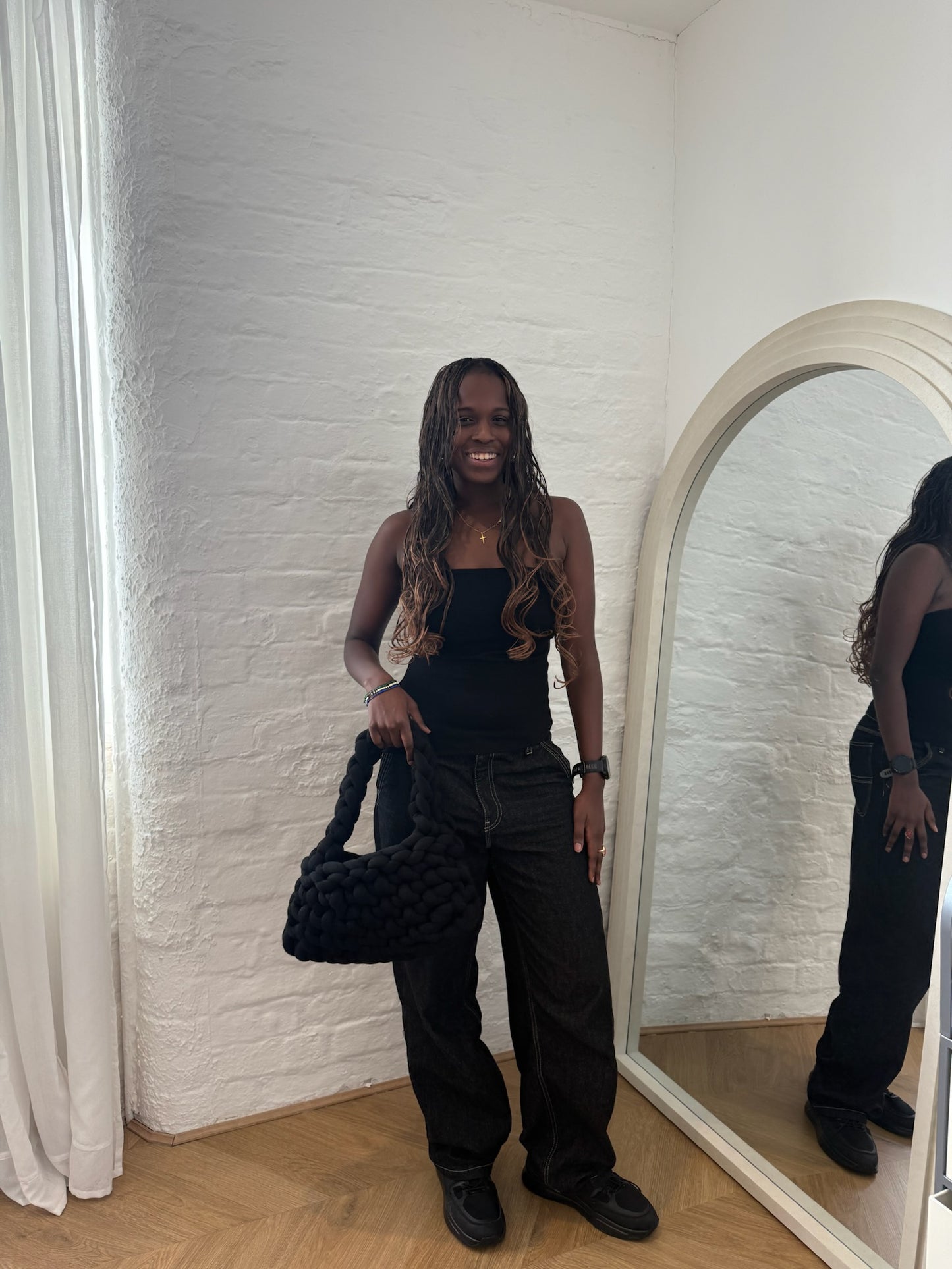 Person standing in front of a mirror holding a black handbag, wearing a black outfit.